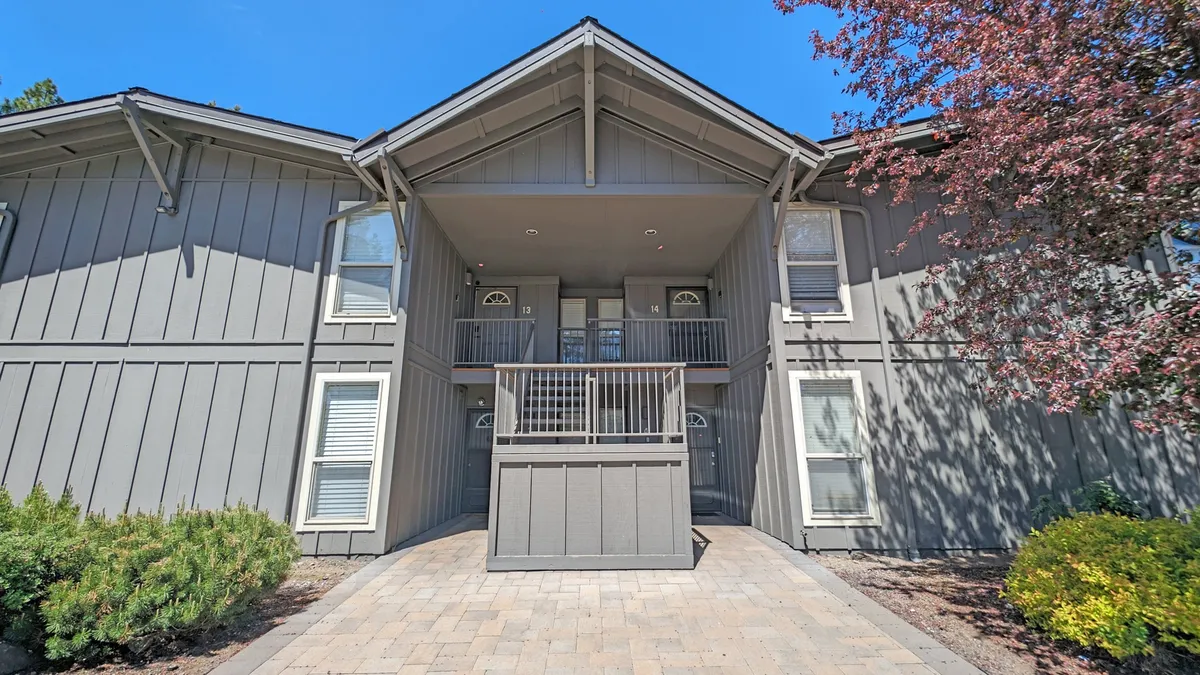 The entrance porch of a condo complex, with a balcony and landscaped shrubs, set among mature trees. The porch and balcony suggest welcoming outdoor spaces ideal for socializing, relaxing, or simply enjoying the view. The landscaping enhances the setting's sense of openness and connection to the outdoors.