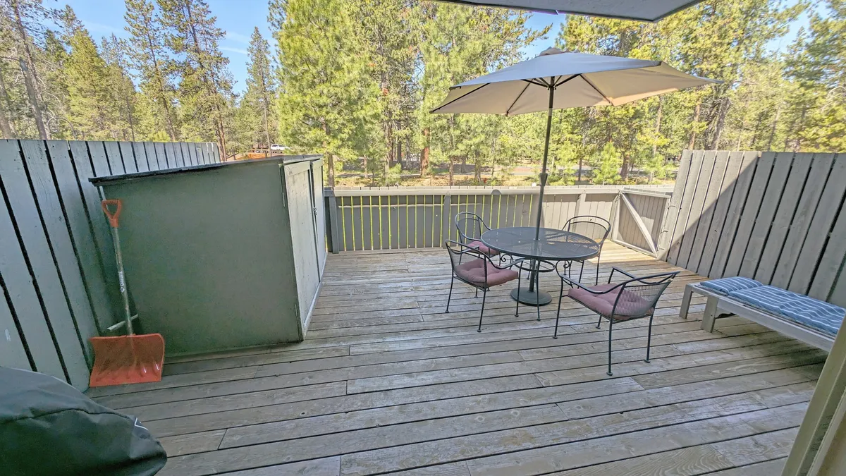 A spacious wooden deck surrounded by tall trees, featuring a table with chairs and a large umbrella for shade. The setting suggests a serene connection to nature and a comfortable spot for outdoor dining or relaxing. The deck overlooks a green, leafy backdrop, inviting guests and their pets to step outside and enjoy the fresh air.