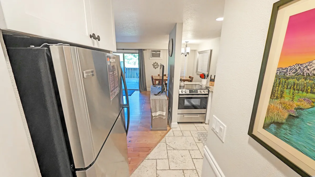 A view into a modern kitchen with stainless steel appliances, a wooden dining table, and large windows letting in natural light and offering an outdoor view. The arrangement invites guests to cook, dine, and converse seamlessly, while the visible artwork and layout support a welcoming mood.