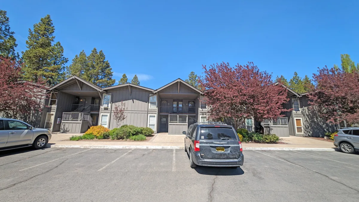 A two-story building surrounded by trees, a parking lot, and flowering shrubs, with open areas suitable for walking and relaxing. The landscaping and natural greenery provide a sense of space and opportunity for outdoor adventures with pets.