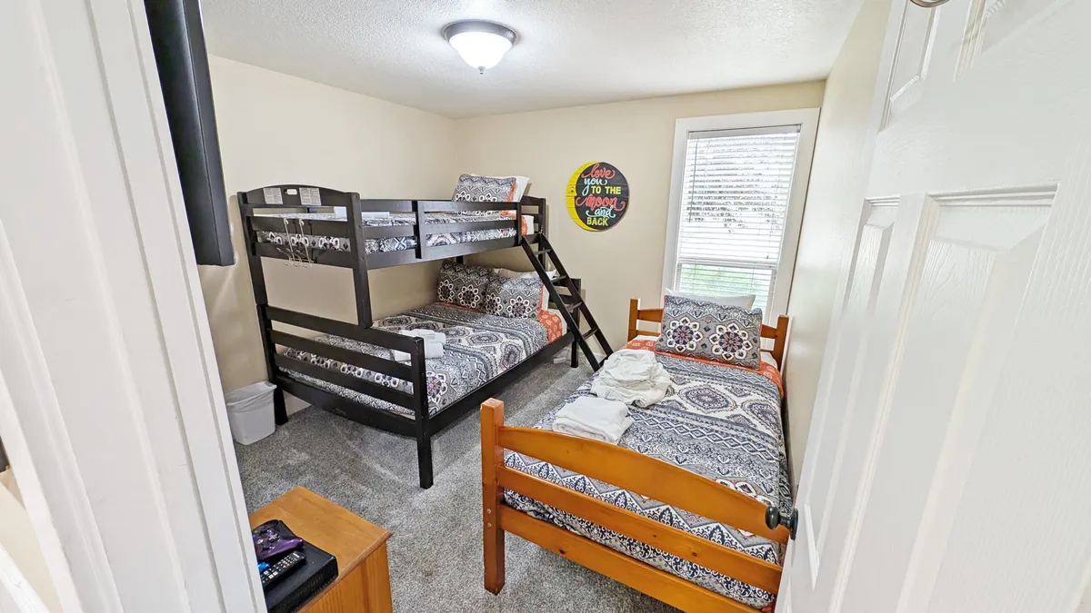 A cheerful and sunny kids' bedroom featuring a sturdy bunk bed and a single bed, playful wall decor, and a window letting in natural light. The space is bright and inviting, with cozy bedding and a relaxed, playful atmosphere.