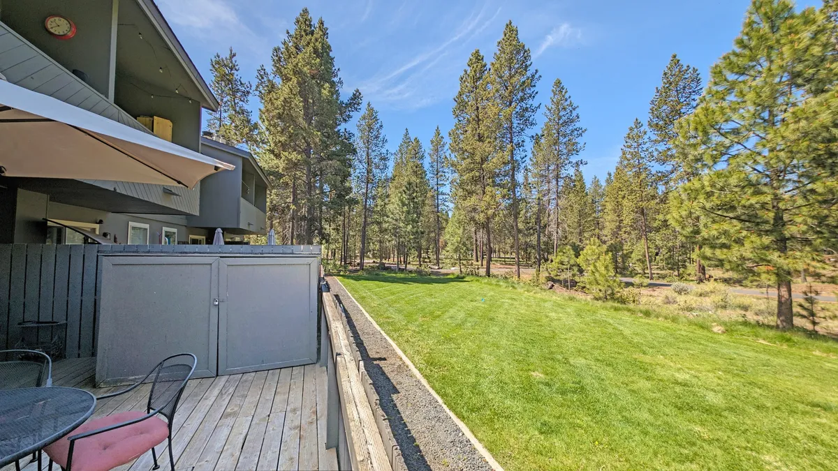 This exterior image features a green lawn bordered by tall pine trees, with patio furniture set up for outdoor dining or relaxing. The natural landscape evokes a peaceful, pet-friendly area for walks and play, extending the sense of space beyond the private deck.
