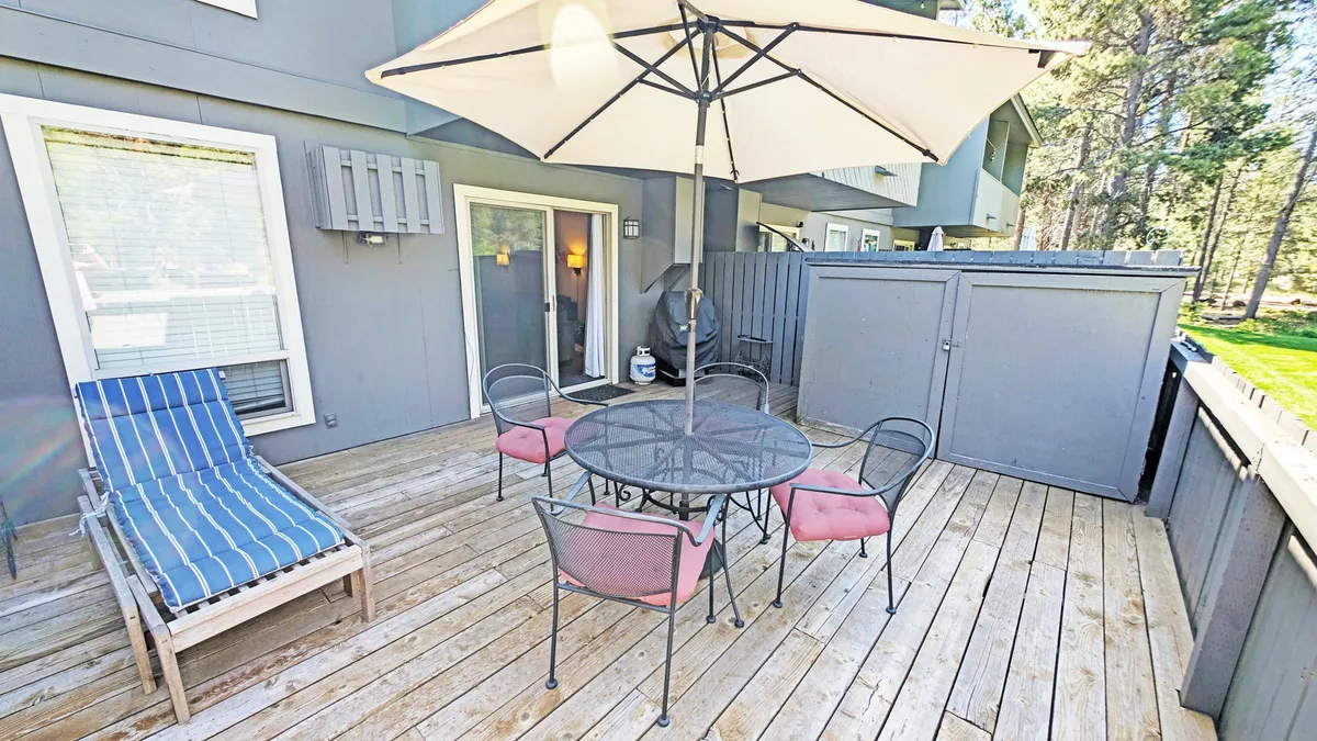 This exterior image showcases a wooden deck featuring an outdoor dining table, a lounge chair, and a large umbrella, with the manicured green space and trees visible beyond. The deck is sunlit and inviting, illustrating the ideal spot for al fresco dining, relaxation, and enjoying the outdoors.