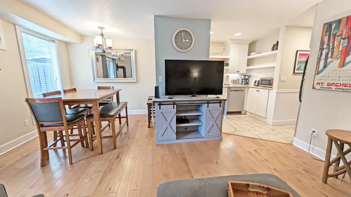 A warm interior featuring a dining area with a wooden table, a television in view, and a mirror that expands the sense of space. The proximity of the kitchen area highlights the open-plan layout, ideal for family meals, game nights, and entertainment.