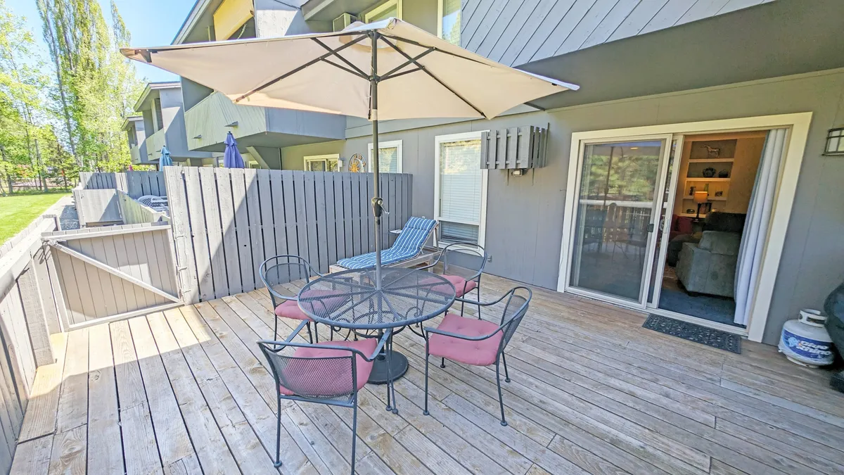 This photo presents a round outdoor dining table with chairs and a patio umbrella on a wooden deck, adjacent to a sliding glass door, suggesting easy indoor-outdoor flow. The setting feels casual and sociable, perfect for meals, gatherings, or a quiet moment with a pet nearby.