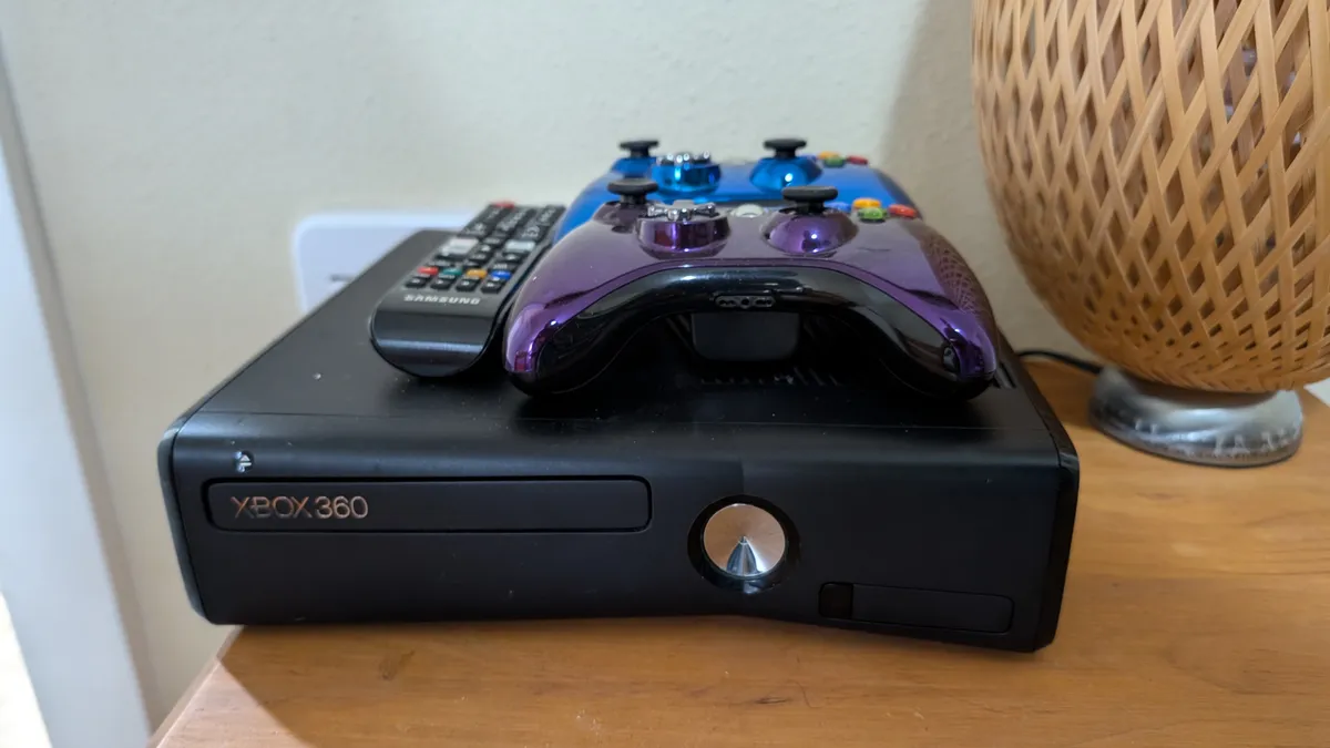 A close-up of an entertainment center with an Xbox 360 console, purple game controller, and remote control, complemented by a stylish woven lamp. The scene suggests a space designed for gaming and movie nights.
